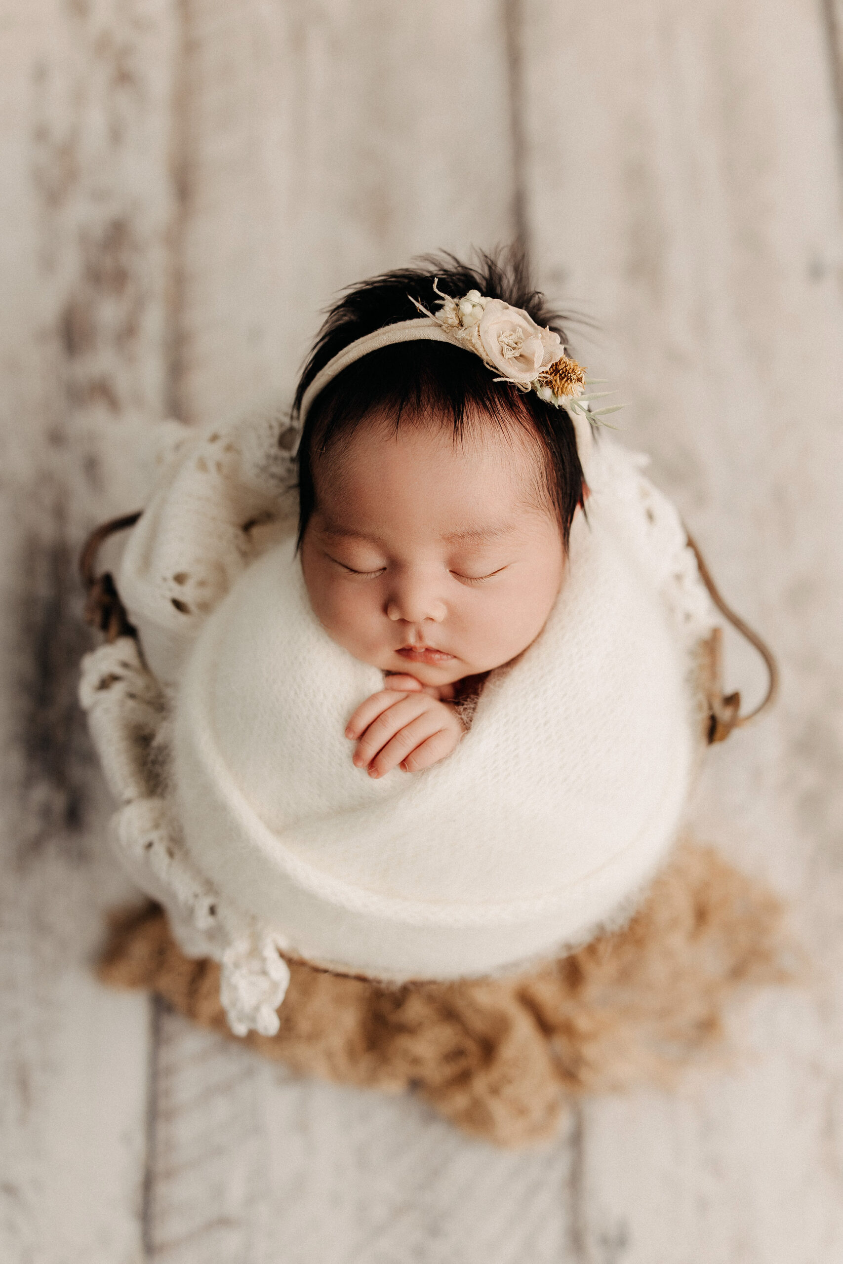 San Diego newborn photography – sleeping baby girl wrapped in creamy white blanket with floral headband, posed in a basket on rustic wood floor